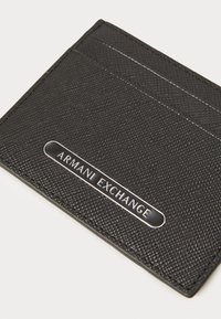 Juodas tekstūruotas kortelių laikiklis, pasižymintis sidabriniu "ARMANI EXCHANGE" logotipu priekyje, su trimis kortelių lizdais, matomais viršuje.