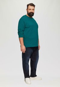 Türkises Langarmshirt mit strukturellen Kontrasten, kombiniert mit dunklen Jeans und weißen Sneakers. Schlichte, lässige Passform und minimalistische Designdetails.