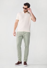 Polo à manches courtes crème avec un col en v ; associé à un pantalon vert clair et des chaussures brunes à enfiler. Les accessoires comprennent des lunettes de soleil noires.