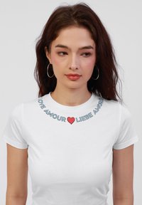 Hvid kortærmet t-shirt med et grafisk tekstdesign, der viser ordene "LOVE AMOUR ❤️ LIEBE AMORE" i blå og rød. Blødt bomuldsstof.