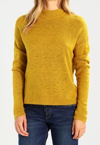 Suéter amarillo de punto con mangas largas y cuello alto. La tela tiene un acabado ligeramente texturizado y un ajuste holgado. Se lleva con jeans azules.