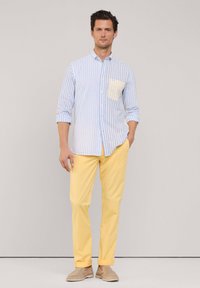 Camisa de botones a rayas azul claro y blanca con un bolsillo amarillo, combinada con pantalones amarillos y zapatos de lona beige. Textura de tejido suave.