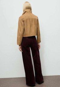 Femme aux longs cheveux blonds portant une veste courte en daim beige et un pantalon large taille haute bordeaux foncé, face à un mur uni.