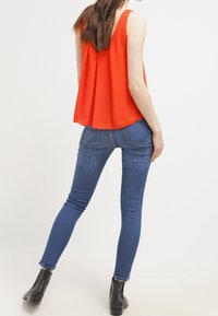Červený top bez rukávů s volným střihem a skládáním na zádech, kombinovaný s přiléhavými modrými džíny a černými kotníkovými botami.
