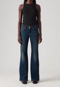 Femme portant un jean évasé foncé, un haut noir sans manches côtelé et des chaussures noires à bout carré, debout devant un fond uni.