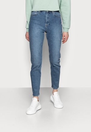 Persoon draagt blauwe slim-fit jeans, witte sneakers en een lichtgroene cropped trui, staand tegen een effen achtergrond.