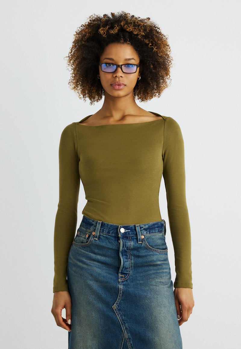 Femme aux cheveux bouclés portant des lunettes à verres bleutés, un haut à manches longues vert olive et une jupe en jean bleu taille haute, tournée vers l'avant.