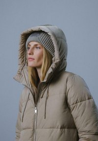Beżowa ocieplana kurtka z wysokim kapturem, z zamkiem błyskawicznym z przodu i sznurkami do regulacji. W zestawieniu z jasnoszarym, prążkowanym beaniem.