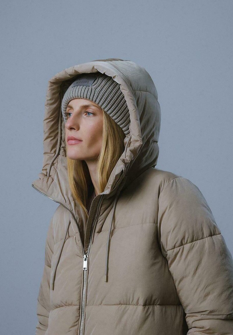Beżowa ocieplana kurtka z wysokim kapturem, z zamkiem błyskawicznym z przodu i sznurkami do regulacji. W zestawieniu z jasnoszarym, prążkowanym beaniem.