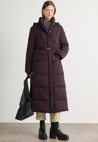 Lang, puffer frakk i mørk lilla med hætte, med quiltet design og belted talje. Sammen med beige bukser og sorte, kraftige støvler.