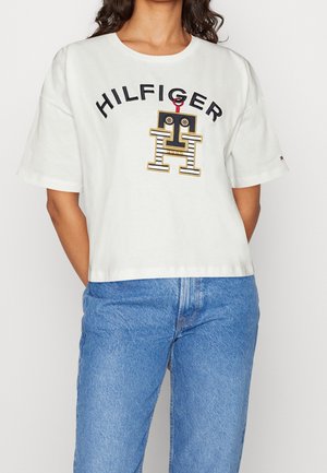 Kobieta ubrana w biały krótki T-shirt z napisem "HILFIGER" i logo w postaci paskowanego robota, w połączeniu z wysokim stanem jasnoniebieskich dżinsów.