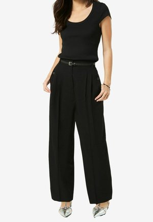 Vrouw draagt een zwart topje met korte mouwen, ingestopt in een hoge taille, wijde zwarte broek met ceintuur en zilveren pumps met puntige neus.