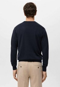 Granatowy sweter z długimi rękawami, okrągłym dekoltem i ściągaczami na mankietach. Materiał wydaje się miękki; noszony z jasno-beżowymi spodniami z tylnymi kieszeniami.