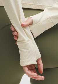 Top crem cu mânecă lungă, cu un model texturat de herringbone, manșete ajustate și orificii pentru degete, purtat cu colanți de culoare verde măsliniu.