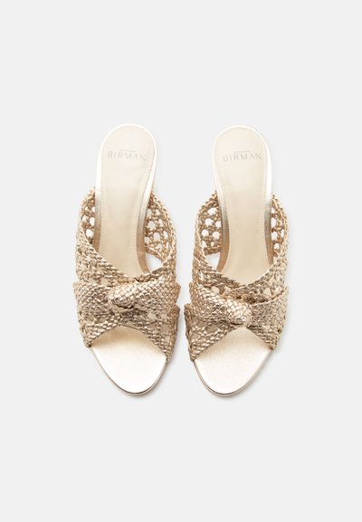 Ζευγάρι χρυσά πλεκτά σανδάλια slip-on με δεμένες λουριά και επωνυμα ενδείκτες σε μονόχρωμο φόντο.