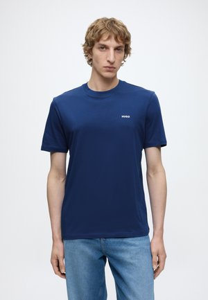 Jeune homme aux cheveux bouclés portant un t-shirt Hugo bleu uni et un jean bleu clair sur fond blanc.