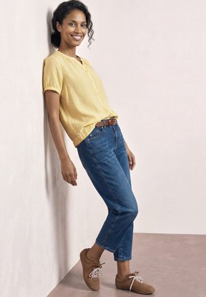 Femme souriante appuyée contre un mur portant une blouse jaune, un jean bleu, une ceinture marron et des baskets marron avec les chevilles croisées.