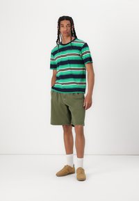 Gestreiftes T-Shirt in Grün, Blau und Marineblau mit kontrastierenden weißen und braunen Streifen; kombiniert mit olivgrünen Shorts und beigen Turnschuhen.