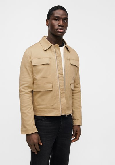 Μπεζ, καπιτονέ μπουφάν με φερμουάρ μπροστά και τέσσερις τσέπες με μπαλώματα. Κατασκευασμένο από απαλό ύφασμα, διαθέτει γιακά και ίσια κοπή.