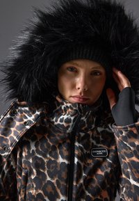 Leopardmønstret jakke med sort faux fur-hette, lynlås-lukning og sorte detaljer. Har et mærket patch på brystet og fingerløse handsker.