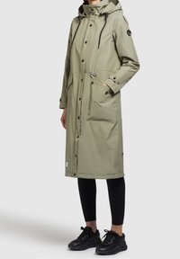 Parka longue vert olive avec capuche, cordons de serrage noirs, boutons-pression, deux poches avant, et un patch logo sur la manche. Portée avec des leggings noirs.
