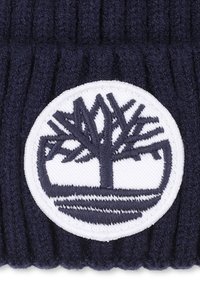 Bonnet tricoté bleu marine avec un patch rond blanc présentant un logo d'arbre bleu foncé. Sa texture côtelée et son matériau doux offrent de la chaleur.