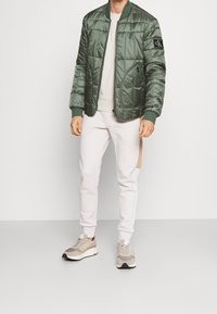 Grøn polstret bomberjakke med ribbede manchetter, lys grå top, hvide sweatpants med sideliste og beige sneakers med teksturerede detaljer.