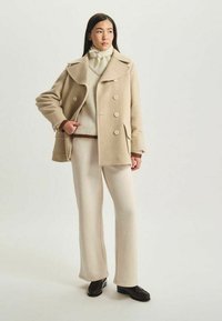 Manteau en mélange de laine beige avec de grands boutons, associé à un pull en tricot crème et un pantalon large assorti. Des chaussures noires complètent la tenue.