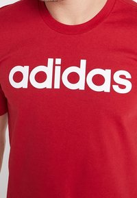 Muž v červeném tričku s bílým logem "adidas" tištěným přes hrudník.