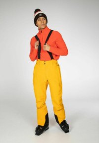 Heldere oranje lange mouw top onder zwarte bretels, gecombineerd met gele skibroek met zwarte accenten bij de enkels en zwarte skischoenen.