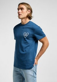 Marinblå bomulls T-shirt med ett hjärtformat logotyp med texten "1889 LEE" i vitt, korta ärmar och rund hals. Slät textur.