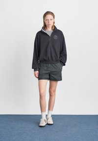 Zwarte sweatshirt met een hoge kraag en rits, gecombineerd met donkere shorts. Het materiaal lijkt zacht; de outfit heeft een subtiele logodetail.