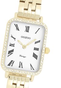 Montre rectangulaire dorée avec un cadran orné de cristaux, cadran blanc, chiffres romains noirs et aiguilles dorées sur un bracelet en maillons.