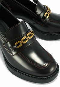 Czarne skórzane mokasyny mają błyszczące wykończenie, burgundowe przeszycia oraz złoty łańcuszek na przodzie. Smukły, zaokrąglony design noska.