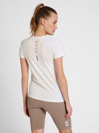 T-shirt blanche à manches courtes avec un design au dos comportant des flèches pêche et une ligne noire verticale. Associée à un short de vélo marron.