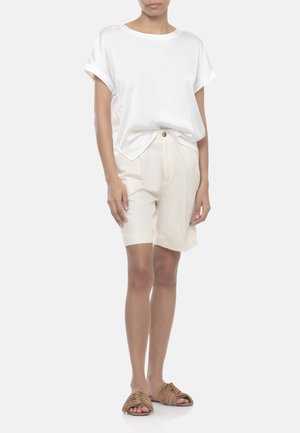 Donna che indossa una camicia bianca a maniche corte larga, pantaloncini beige fino al ginocchio e sandali marroni con frange, in piedi con postura rilassata.