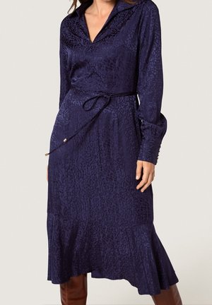 Femme portant une robe bleu marine à manches longues, longueur genou, à imprimé léopard subtil, avec un décolleté en V, une ceinture à nouer à la taille et un ourlet volanté.