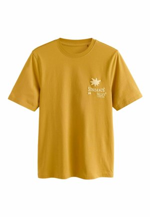 Młody mężczyzna w musztardowożółtej koszulce z napisem "Sundance Valley 1999" i grafiką słońca, patrzy w lewo na neutralnym tle.