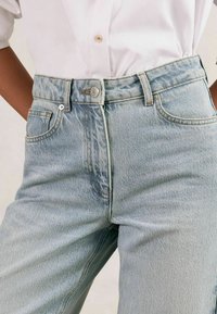 Gai zilu denima bikses ar augstu jostasvietu, priekšējiem kabatām un metāla pogas aizdari. Iezīmējas ar izbalējušu tekstūru un kontrastējošām šuvēm.