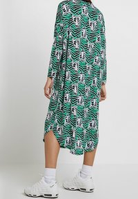 Kvinna som bär en midi-klänning med tigertryck i grönt, svart och vitt, långa ärmar, vita ankelsockor och vita sneakers, ståendes på en enfärgad bakgrund.