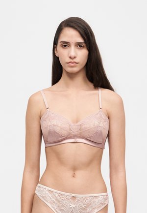 Une femme porte un soutien-gorge en dentelle rose pâle et une culotte assortie sur un fond blanc uni.