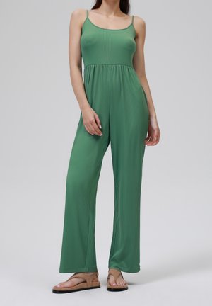 Zielony kombinezon bez rękawów z dopasowaną górą i szerokimi nogawkami. Wykonany z miękkiego, gładkiego materiału o płynnej fakturze. Noszony z jasnobrązowymi sandałami.