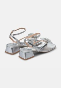 Zilveren metallic sandalen met een blokhak, voorzien van kruislings gespen en een structuuraccent aan de voorkant. Glad interieur en een gespsluiting.