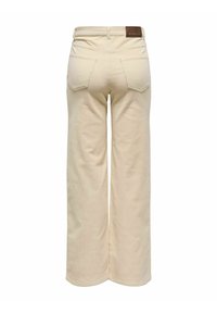 Pantalons larges beiges en tissu texturé, avec deux poches arrière et une étiquette de marque en cuir visible à la taille.