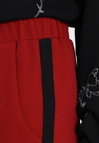Rode stoffen broek met een geplooid ontwerp en zwarte accenten aan de zijkanten, gecombineerd met een zwart top met witte bloemenborduursels.
