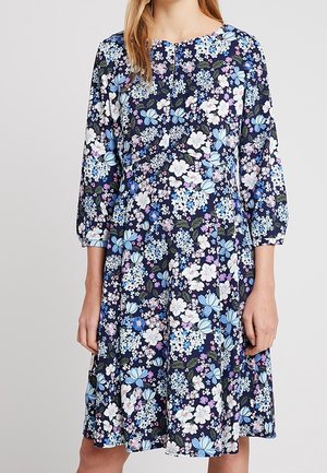Femme portant une robe bleu marine longueur genou avec des manches trois-quarts et un motif floral multicolore en tons blancs, bleus et violets.