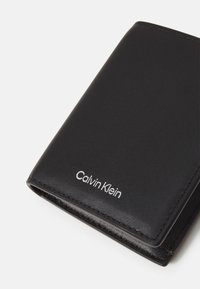Svart lærpung med en glatt tekstur, med "Calvin Klein"-logoen i hvitt i nederste hjørne. Elegant, minimalistisk design.