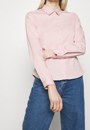 Personne portant une chemise rose clair à manches longues avec col et boutons, et un jean bleu, debout devant un fond blanc uni.