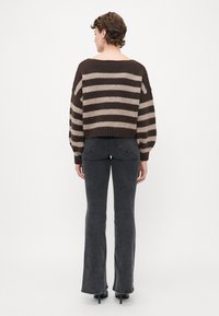Woman facing away wearing a brown and beige striped sweater, dark gray flared jeans, and black heeled shoes against a plain white background.