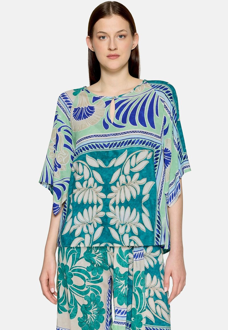 Femme portant un haut ample et un pantalon assortis avec des motifs floraux et géométriques verts, bleus et beiges, debout devant un fond uni.